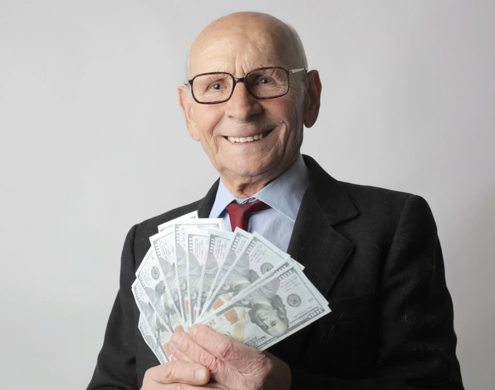 Smiling elderly man in a suit and tie, holding a fan of dollar bills, exuding happiness and financial success