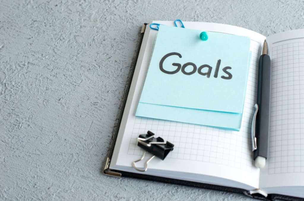 "Goals" written on a sticky note clipped in a checkered notebook with a pen
