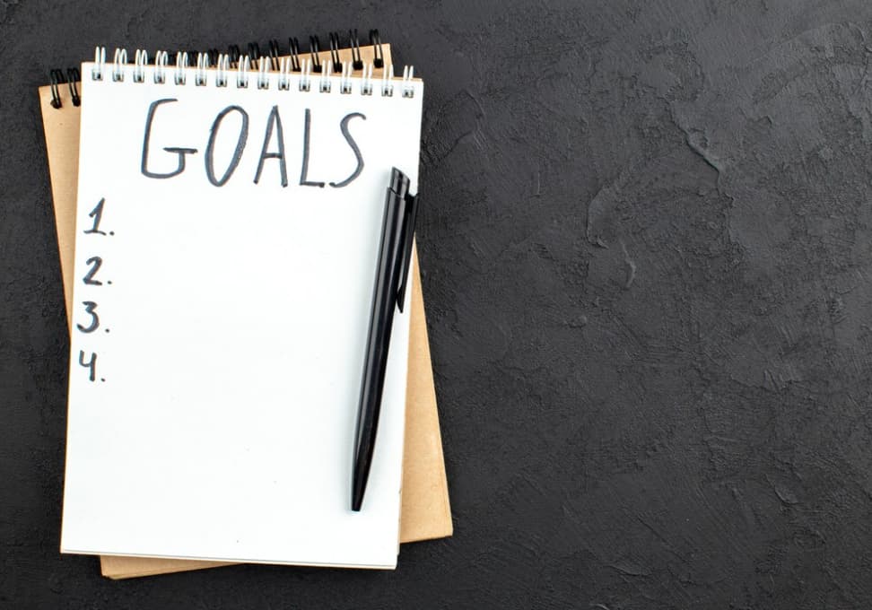 Notepad with "GOALS" at the top and a numbered list, beside a pen, on a dark background